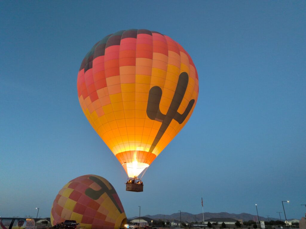 Hot-air balloon glowing from its burner while airborne over the West Valley before sunrise, with another balloon below.