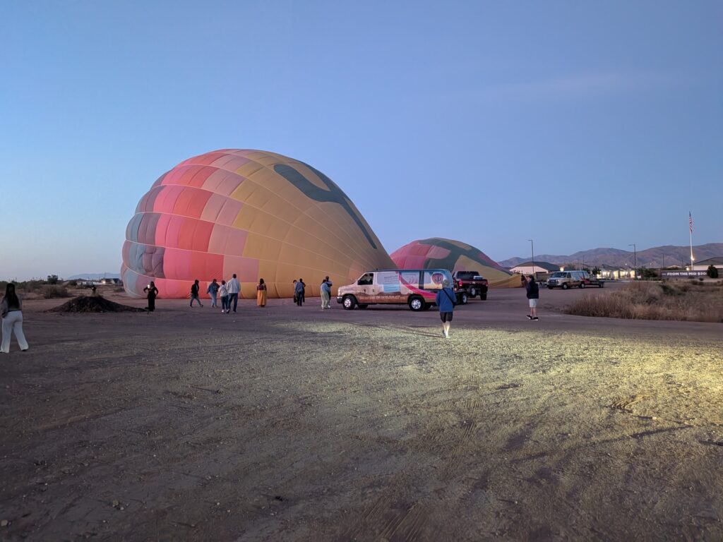 Hot-air balloons laid out and being prepared for inflation before sunrise, with passengers and support vehicles nearby.