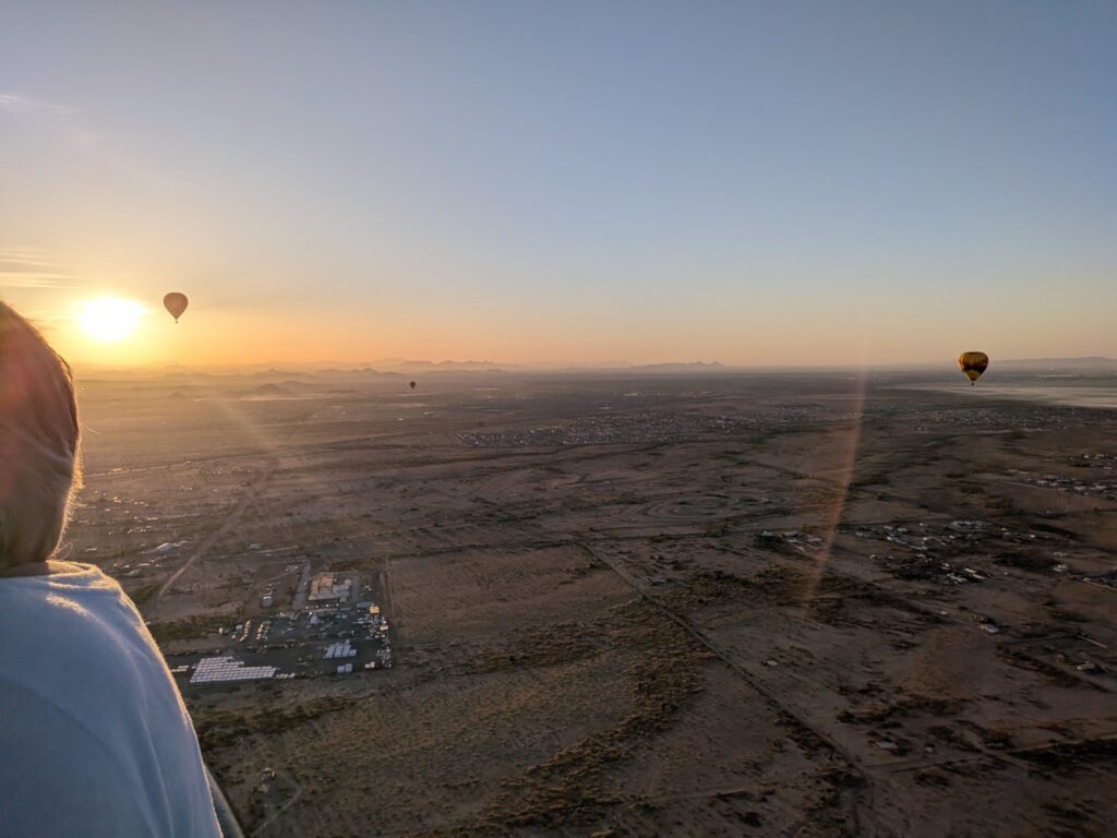 View from a hot-air balloon over the West Valley at sunrise, with several other balloons visible in the distance.